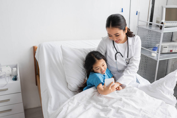carefree child taking selfie on smartphone with smiling asian doctor in hospital ward - Foto, Imagem