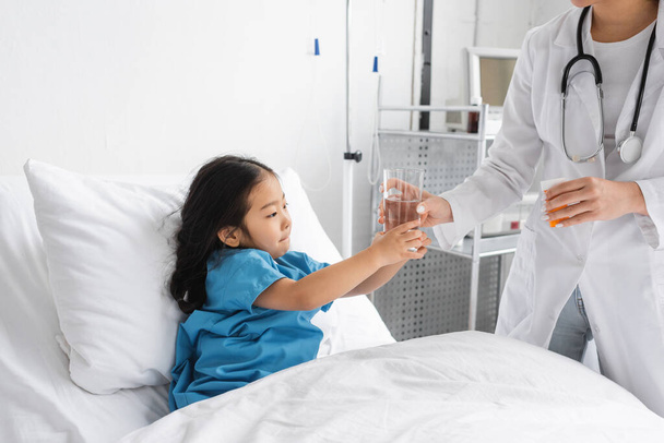 白衣を着た医者が薬の容器を持って病院のベッドの上のアジアの女の子に水を与え - 写真・画像