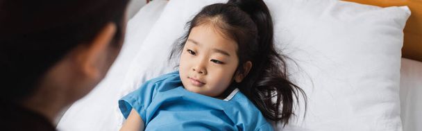 diseased asian girl looking at blurred pediatrician while lying on hospital bed, banner - 写真・画像