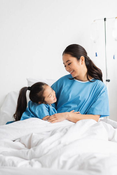 happy asian mother and daughter embracing and looking at each other on hospital bed - Фото, зображення