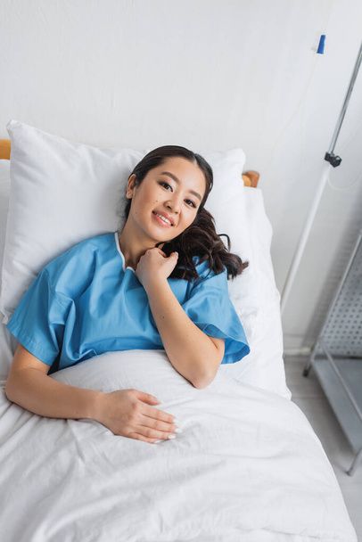 Hochwinkelaufnahme einer zufriedenen asiatischen Frau, die auf dem Krankenhausbett liegt und in die Kamera blickt - Foto, Bild