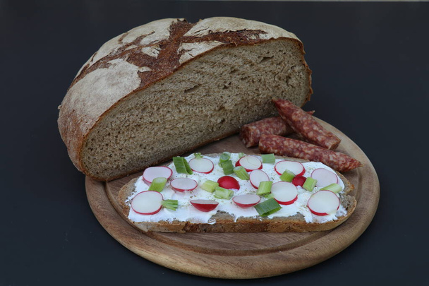 une demi-miche de pain cuit en pierre avec un sandwich traditionnel Pajottenland d'une tranche de pain tartiné de fromage frais saupoudré de radis et d'oignons de printemps et de saucisses séchées sur le côté - Photo, image