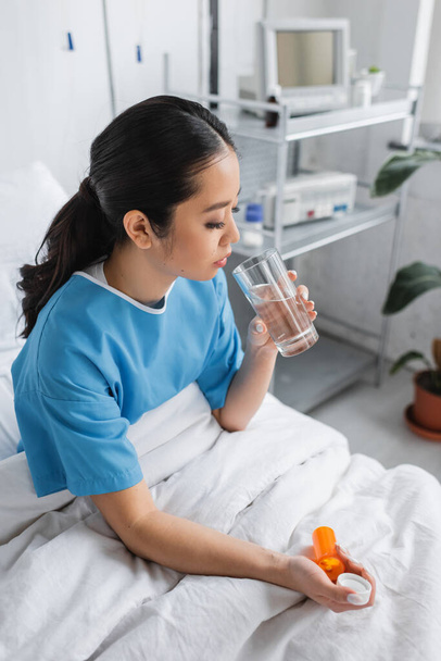 брюнетка азіатська жінка в лікарняній сукні сидить на ліжку зі склянкою води і таблетки контейнер
 - Фото, зображення