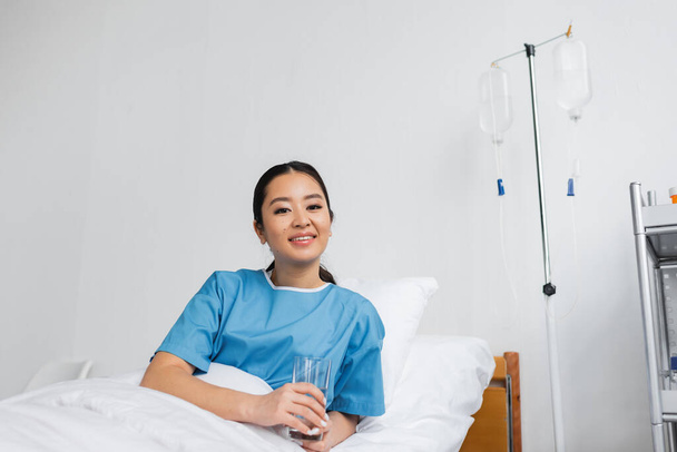 веселая азиатская женщина держит стакан воды и смотрит в камеру на больничной койке - Фото, изображение