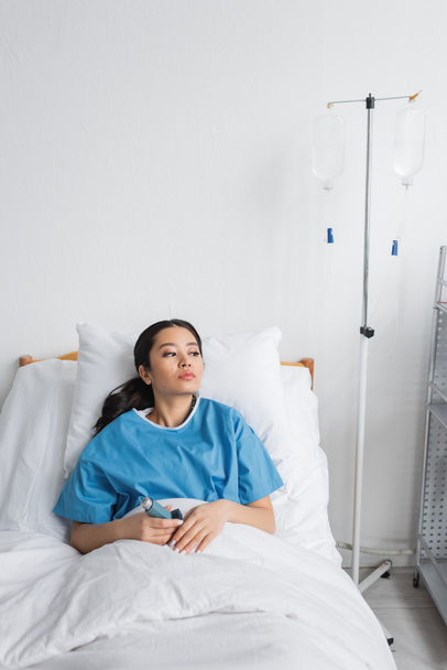 přemýšlivá asijská žena drží inhalátor a dívá se jinam na lůžko v nemocnici oddělení - Fotografie, Obrázek