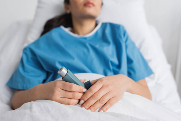 bijgesneden zicht op wazige vrouw die inhalator vasthoudt terwijl ze op bed ligt in de kliniek - Foto, afbeelding