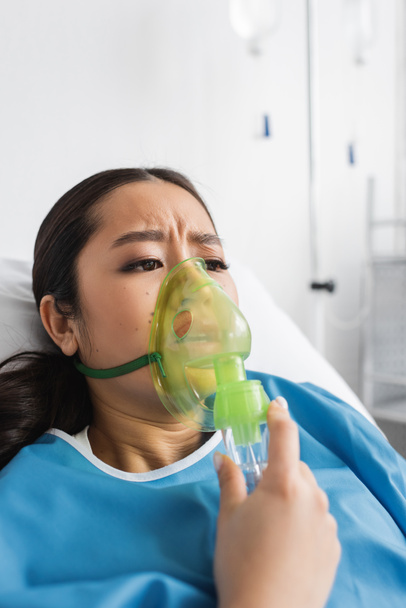 ανησυχούσα Ασιάτισσα που ανέπνεε μάσκα οξυγόνου ενώ ήταν ξαπλωμένη στο κρεβάτι στην κλινική - Φωτογραφία, εικόνα