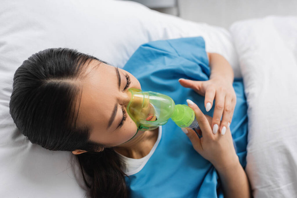 bovenaanzicht van zieke aziatische vrouw in zuurstof masker liggend op bed in ziekenhuis afdeling - Foto, afbeelding