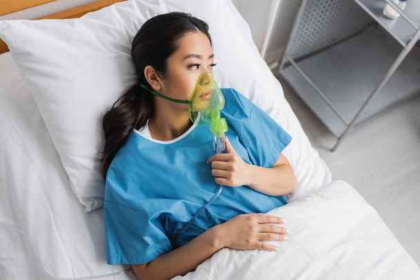 κορυφαία άποψη της αναστατωμένης Ασιάτισσας ξαπλωμένης στο κρεβάτι του νοσοκομείου με μάσκα οξυγόνου και κοιτάζοντας αλλού - Φωτογραφία, εικόνα