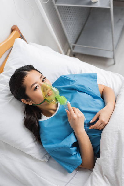 top view of positive asian woman breathing in oxygen mask and looking at camera on bed in clinic - Photo, Image