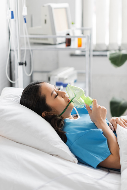 πλαϊνή όψη μιας άρρωστης Ασιάτισσας που κρατάει μάσκα οξυγόνου ενώ είναι ξαπλωμένη στο κρεβάτι στο θάλαμο του νοσοκομείου - Φωτογραφία, εικόνα