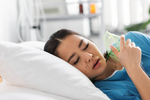 nemocná asijská žena se zavřenýma očima ležící na nemocničním lůžku s kyslíkovou maskou - Fotografie, Obrázek