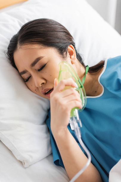 nemocná asijská žena držící kyslíkovou masku při ležení na nemocniční posteli se zavřenýma očima - Fotografie, Obrázek