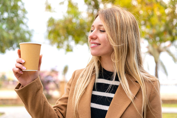Junge hübsche blonde Frau hält draußen mit glücklichem Gesichtsausdruck einen Kaffee zum Mitnehmen - Foto, Bild