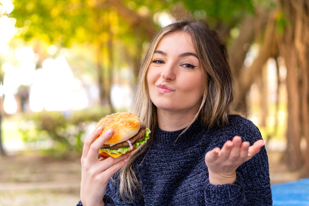 Joven mujer rumana bonita sosteniendo una hamburguesa al aire libre haciendo gesto dudas mientras levanta los hombros - Foto, imagen