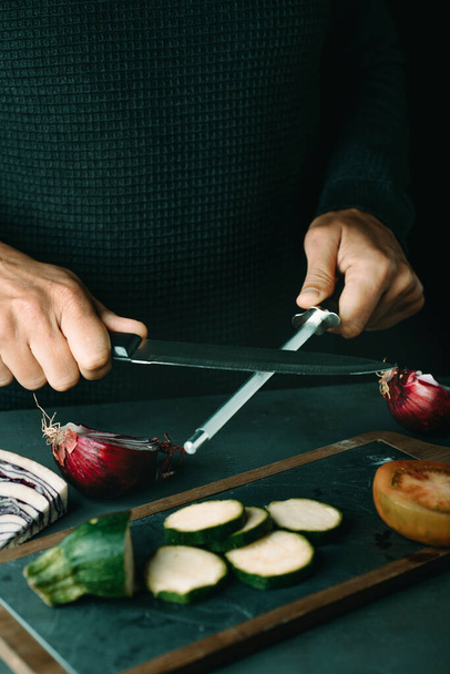 man steeling a kitchen knife with a honing steel at a table with some chopped vegetables - Photo, Image