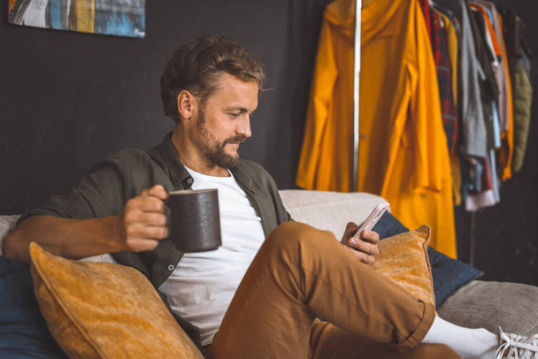 Hombre tomando un descanso en casa, disfrutando de una taza de café mientras se concentra en el teléfono. Se enfatiza el concepto de pasar tiempo en casa, destacando la importancia de la relajación y el ocio en nuestras rutinas diarias. - Foto, imagen
