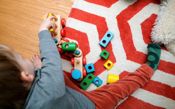 Un bébé gai joue avec des jouets en bois colorés dans une chambre d'enfant confortable, couché sur le sol entouré de jouets colorés de différentes formes - Photo, image