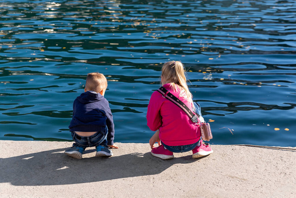 Back view two cute little curious sibling kids sitting on concrete embankment pier near sea lake bank and watching fish ship yacht boat marina. Children enjoy have fun playing outdoors at summer day. - Foto, imagen
