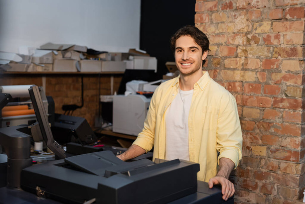 szczęśliwy człowiek w żółtej koszuli stoi w pobliżu profesjonalnego sprzętu w centrum druku - Zdjęcie, obraz