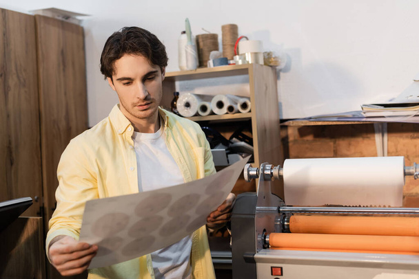 tipógrafo mirando papel impreso cerca de plotter de impresión profesional - Foto, Imagen