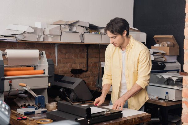 tipógrafo de buen aspecto corte de papel con cuchillo cerca de recortadora de papel en el centro de impresión  - Foto, Imagen
