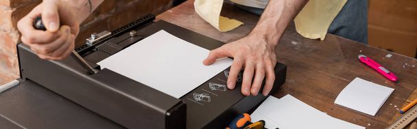 vista recortada del hombre tatuado utilizando la máquina cortadora de papel en el centro de impresión moderno, pancarta  - Foto, Imagen