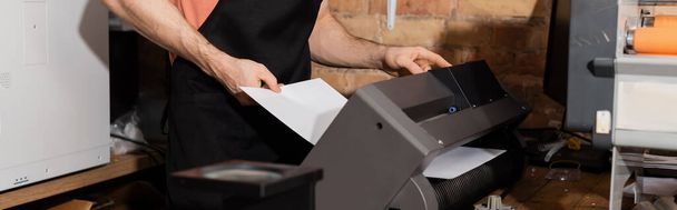 vista cortada do trabalhador em avental segurando papel perto de impressora e jornais frescos no centro de impressão, banner  - Foto, Imagem
