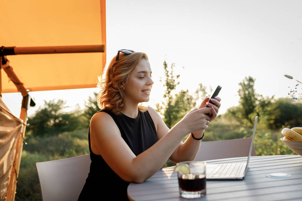 Smiling Woman freelancer using phone while working on laptop in cozy glamping tent in a sunny day. Luxury camping tent for outdoor summer holiday and vacation. Lifestyle concept - Foto, Bild