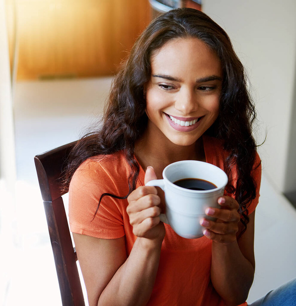 Beginning her day with a fresh cup of coffee. a smiling young woman drinking coffee while sitting in a chair at home - 写真・画像