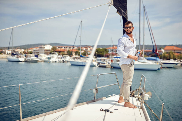 A young man standing on the bow of the yacht and enjoying the view on the town on the seaside on a beautiful sunny day. Summer, sea, vacation - Fotografie, Obrázek