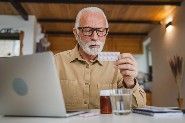 Jeden senior mężczyzna kaukaski mężczyzna dziadek siedzieć w domu trzymać blister tabletki przeciwbólowe lub witaminy suplement czytać etykieta gotowy do podjęcia leków w pobliżu laptop zamówić online przestrzeń kopia - Zdjęcie, obraz