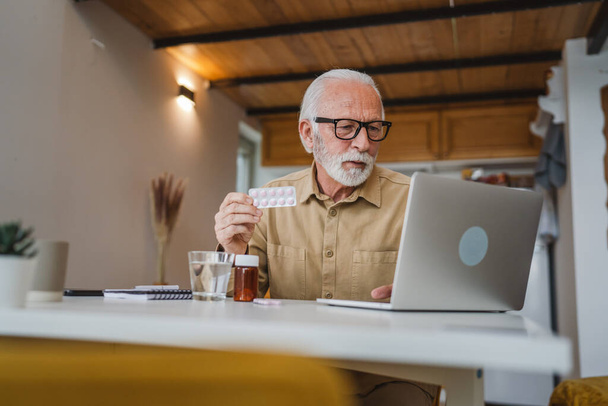 Yksi vanhempi mies valkoihoinen mies isoisä istuu kotona pidä läpipainopakkaus huumeiden tabletti kipulääkkeitä tai vitamiini täydentää lue etiketti valmis ottamaan lääkettä lähellä kannettavaa tietokonetta järjestyksessä verkossa kopioi tilaa - Valokuva, kuva