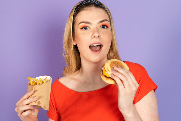 Opgewonden verrast knappe meid eet fastfood, goedkope hamburger, friet en hamburger, open mond. fastfood en ongezond dieet. geïsoleerd op paarse achtergrond. - Foto, afbeelding