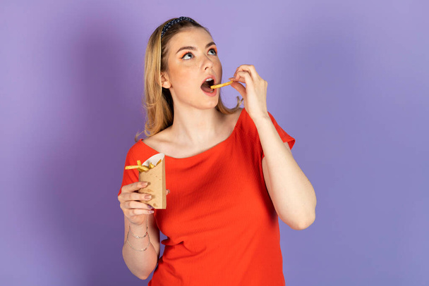 Opgewonden hongerig mooi meisje eet fast food, olieachtig geprobeerd aardappel, met frietjes in haar hand. fastfood en ongezond dieet. geïsoleerd op paarse achtergrond. - Foto, afbeelding