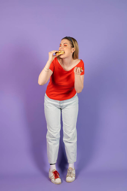 In Mayonnaise geschmiert isst hübsches Mädchen Fast Food, einen billigen Burger, hält Cheeseburger in der Hand. Fast Food und ungesunde Ernährung. isoliert auf violettem Hintergrund. - Foto, Bild