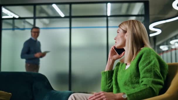 Smiling businesswoman greeting colleague at hall. Positive woman talking smartphone sitting armchair at lounge closeup. Man associate waving to calling lady at lobby zone. Corporate lifestyle concept - Footage, Video