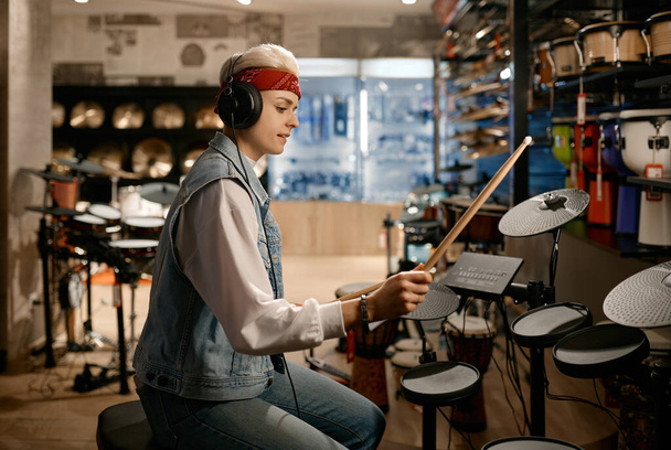 Молодая хипстерша-музыкантка в наушниках играет на электронном барабане в магазине музыкальных инструментов - Фото, изображение