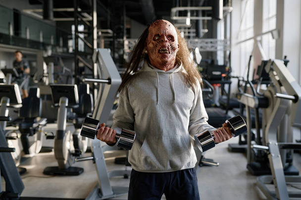 Zombie man z przerażającą twarzą ćwiczy z hantlami trenując mięśnie w nowoczesnym klubie sportowym - Zdjęcie, obraz