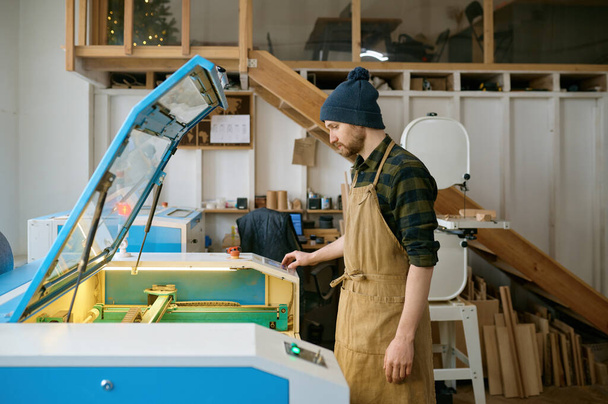 Mann Tischler, der CNC-Lasermaschine im automatisierten Holzbearbeitungsstudio bedient. Fertigungs- und Engineering-Prozess in der Tischlerei - Foto, Bild