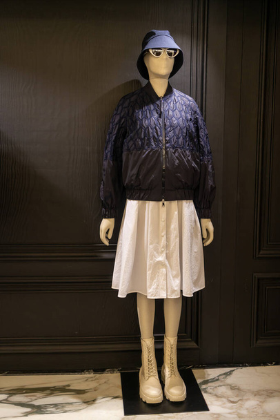 The mannequin is dressed in a dark blue pilot jacket, a white skirt, a panama hat, sunglasses and white high top sneakers against a black wall. - Valokuva, kuva