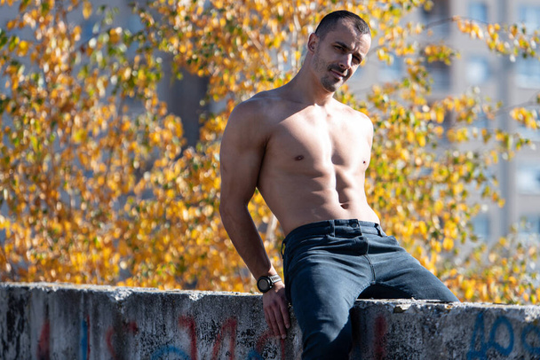 Healthy Young Man Sitting Strong Flexing Muscles on Old Rooftop - Muscular Athletic Bodybuilder Fitness Model Posing Outdoors - Photo, Image