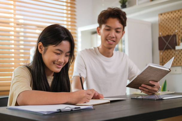 Usmívající se asijské male tutor pomáhá dospívající dívka dělá domácí úkoly na stole doma. - Fotografie, Obrázek