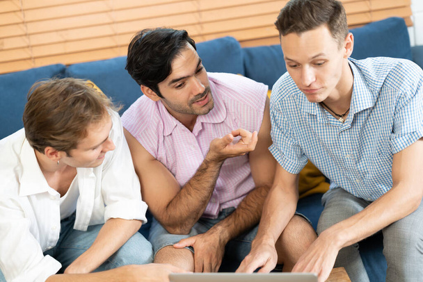 Dreifacher junger Mann, der im Wohnzimmer sitzt, während er mit Laptop Videotelefonie mit Regenbogenfahne auf dem Schreibtisch führt. LGBTQ-Menschen leben und lieben Emotionen. LGBT-Soziales Netzwerk. Selektiver Fokus - Foto, Bild