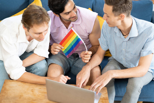 Тройной молодой человек сидит в гостиной, используя ноутбук для видео-звонка с прапорцем веселки на столе. ЛГБТ-люди живуть і люблять емоції. Соціальна мережа ЛГБТ. зосередження уваги - Фото, зображення