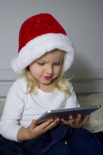 Pieni vaalea tyttö punaisessa Santa hattu istuu sängyllä ja tilalla tabletti, lapsi käyttää internetiä gadget, lapsi riippuvainen tabletin ja etsivät sarjakuvia, lapsi studiyng verkossa kehityshäiriöitä pelejä - Valokuva, kuva