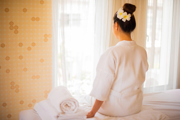 Mooie vrouw in witte badjas Leun achterover op het bed terwijl u kijkt naar het gordijn in de spa kamer om massage voor te bereiden met professionele masseuse. - Foto, afbeelding