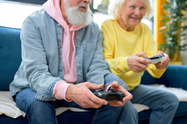 Happy man and woman in casual clothes sit on sofa at cozy room playing video game console, have rest at weekends. Mature people playing entertaining video game battle together, focus on hands - Foto, afbeelding