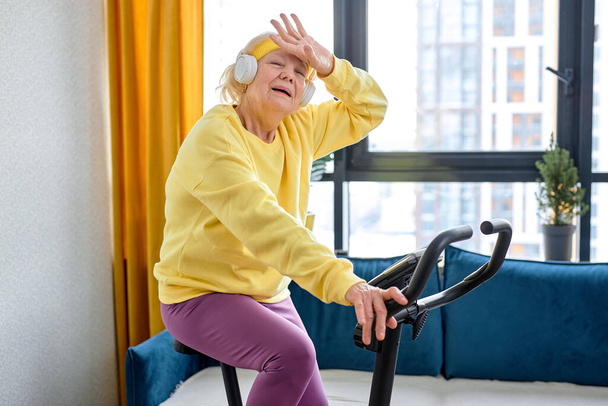 Exhausted person training on stationary bicycle doing physical exercise and activity. Senior caucasian woman using cardio cycling machine to train legs muscles with gymnastics at home, tired - Photo, image