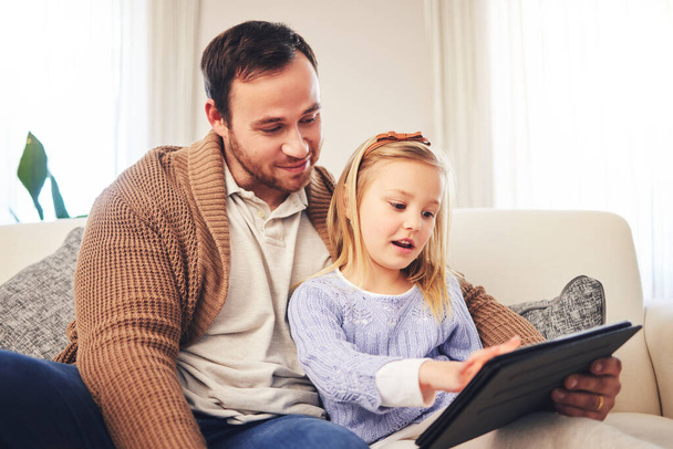 Family, father and a child with a tablet for learning at home while playing a game for education on internet. A man or dad and girl kid on sofa for streaming, mobile app and website for development. - 写真・画像